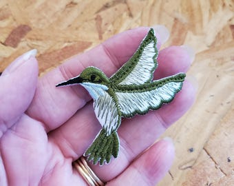 May include: Embroidered hummingbird patch in shades of green, white, and black. The patch features detailed stitching, showcasing the bird's wings, body, and beak. The hummingbird is depicted in mid-flight, with its wings spread. This patch is suitable for clothing or accessories.