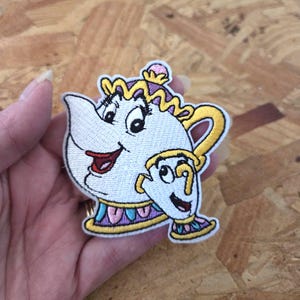 May include: Embroidered patch featuring the characters Mrs. Potts and Chip from Beauty and the Beast. The patch has a white background with yellow, pink, and purple accents. The design is a cartoon style.