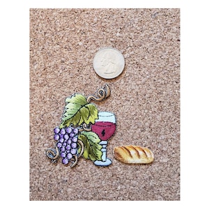 May include: Embroidered patch featuring a wine glass filled with red liquid, a bunch of purple grapes, green leaves, and a loaf of bread. A quarter coin is next to the patch. The patch is on a brown, textured surface.