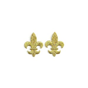 May include: Two gold glitter fleur-de-lis ornaments on a white background. The fleur-de-lis is a stylized lily, a symbol often associated with royalty and heraldry. The ornaments are identical in shape and size.