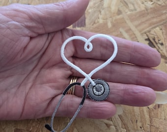 May include: A white and grey embroidered patch shaped like a stethoscope with the tubing forming a heart outline. The chest piece is circular with grey and white stitching, while the ear tubes are outlined in white with grey accents at the ends.