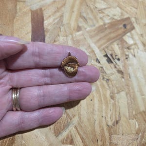 May include: A small, acorn-shaped pendant with a detailed, embroidered design. The pendant is primarily brown with a lighter, tan-colored interior, and is held in a person's hand. The background is a light brown, textured surface.