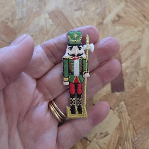 May include: Embroidered patch of a nutcracker soldier in green, red, and gold. The patch features a detailed design of the nutcracker's uniform, hat, and staff. The patch is held in a hand.