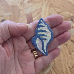 May include: An embroidered patch in the shape of a seashell. The patch features a blue border and blue horizontal stripes, with an iridescent, pearlescent fabric filling the spaces between the stripes, creating a shimmering effect on the surface.