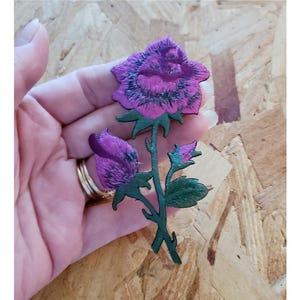 May include: A pink and green embroidered rose patch with a single large rose and two smaller buds. The rose has a dark pink center and a lighter pink outer layer. The leaves are a dark green.