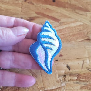 May include: Embroidered seashell patch with a blue outline, white interior, and iridescent pink and gold accents. The patch is in the shape of a conch shell. This decorative patch can be sewn or ironed onto fabric.