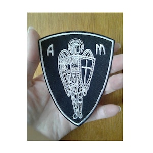 May include: A black and white embroidered patch with a shield and an angel holding a sword. The patch is shaped like a triangle with the letters "A" and "M" on either side of the angel.