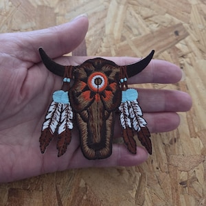 May include: Embroidered patch featuring a brown bull skull with dark horns, an orange and white central design, and blue and white feather accents. The patch is held in a hand.