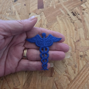May include: A blue embroidered patch featuring a caduceus symbol, which consists of a central staff with two wings at the top and two snakes coiled around the staff in a symmetrical pattern. The patch has a textured, stitched thread finish.