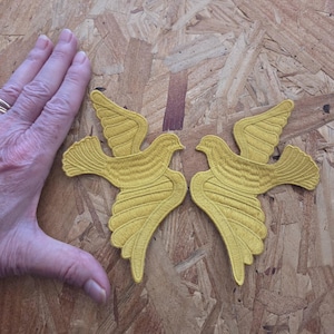 May include: Two mustard yellow embroidered bird patches featuring detailed stitching on the wings and tail feathers. The patches are shaped like birds in flight with outstretched wings, showcasing a consistent thread texture throughout the design.