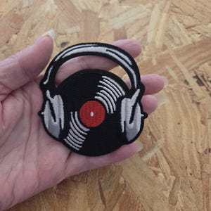 May include: A circular embroidered patch featuring a black vinyl record with a red center label, partially covered by a pair of white and grey headphones. The design uses thick black stitching for the outline and details, creating a high-contrast graphic.