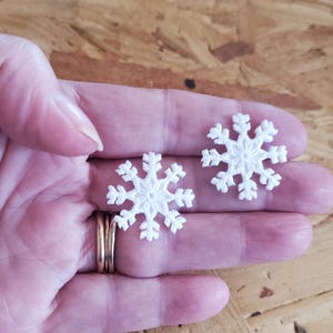 May include: Two white, crocheted snowflake ornaments. Each snowflake has six points and a detailed, openwork design. The snowflakes are held in a hand, showcasing their delicate texture and intricate craftsmanship. The background is a warm, wooden surface.