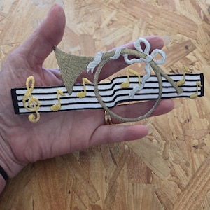 May include: An embroidered patch featuring a gold-colored trumpet with a silver ribbon bow, layered over a horizontal white fabric strip with black horizontal lines and several gold-colored musical notes including a treble clef.
