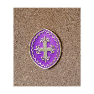 May include: Purple and gold embroidered patch with a cross design. The patch is circular and has a scalloped edge.