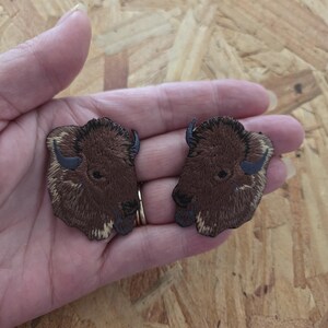 May include: Two embroidered patches featuring a bison head. The patches are brown with dark blue horns and tan accents. The bison heads are facing opposite directions. The patches are held in a hand.