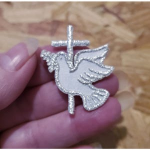 May include: Silver embroidered patch of a dove with outstretched wings, holding a cross in its beak.
