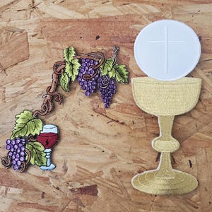 May include: Embroidered patches featuring religious symbols. One patch depicts a chalice with a white wafer and cross, while another shows a grapevine with purple grapes and a glass of red wine. The patches are on a wood-grain background.