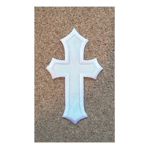 May include: White embroidered cross patch with a simple design. The patch is on a brown corkboard background.