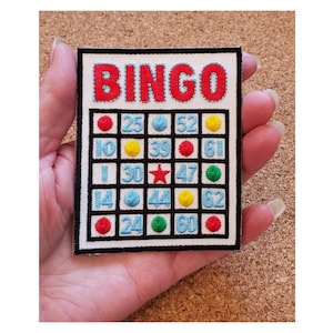 May include: Embroidered patch featuring a bingo card design. The word "BINGO" is in red, above a grid of numbers and colored dots. The patch has a black border and is on a white background.