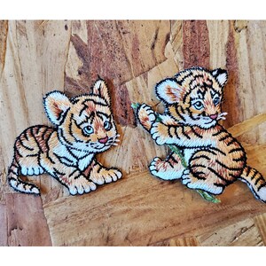 May include: Two embroidered iron-on patches of tiger cubs. One cub is lying down and the other is climbing a green vine.