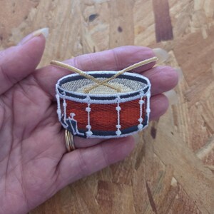 May include: An embroidered patch featuring a red snare drum with white trim and two crossed tan drumsticks resting on the top drumhead. The design includes dark gray hardware details along the sides and base of the drum, rendered in thread.