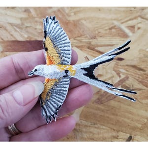 May include: A white, black, and yellow embroidered bird patch with a long tail. The bird is flying with its wings spread out.
