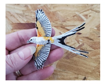 May include: A white, black, and yellow embroidered bird patch with a long tail. The bird is flying with its wings spread out.
