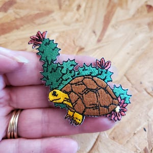 May include: Embroidered patch featuring a turtle with a brown shell, yellow feet and head, and green cactus-like foliage with pink flowers. The patch is designed with detailed stitching and a whimsical aesthetic.