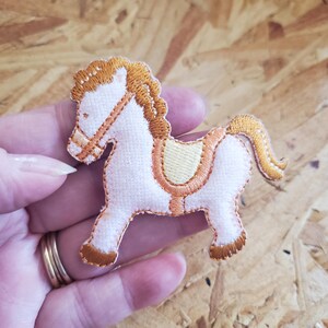 May include: Embroidered patch of a white horse with a tan mane, tail, and saddle. The horse has tan accents on its bridle and hooves. The patch is likely for clothing or accessories.