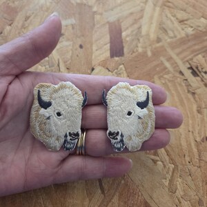 May include: Two embroidered patches featuring a bison head. The patches have a cream-colored background with tan and black details. The bison's horns and facial features are black. The patches are held in a hand.