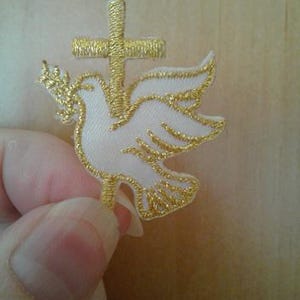 May include: A white dove with gold embroidery, perched on a gold cross. The dove is facing right and has its wings spread. The cross is simple and has a rounded top.