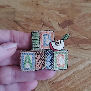 May include: Embroidered patch featuring three colorful alphabet blocks with the letters A, B, and C. A red and white apple slice with a green leaf sits atop the blocks. The patch is a small, handmade item.