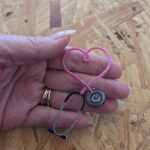 May include: An embroidered patch shaped like a stethoscope with a pink heart-shaped tube and a circular grey chest piece. The design features a black and grey lower section with a pink upper section forming a heart outline at the top.