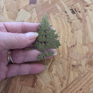 May include: A detailed, green, tree-shaped ornament with a textured surface. The ornament is held against a wood-grain background, showcasing its intricate design. The tree has a delicate, layered appearance, suggesting a handcrafted item.