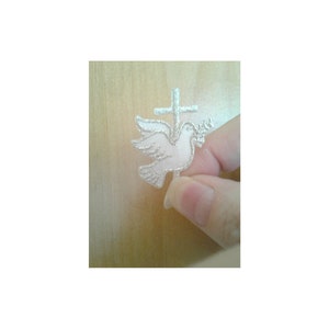 May include: A white dove with an olive branch in its beak perched on a silver cross. The dove and cross are embroidered on a white fabric.