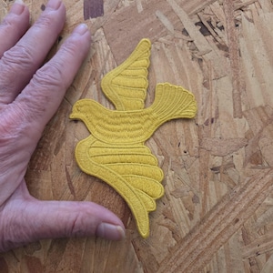 May include: A mustard yellow embroidered patch in the shape of a bird in flight with wings spread wide. The design features detailed stitching lines throughout the body and wings, creating a textured appearance for this fabric applique.