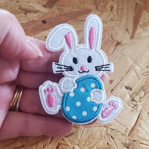 May include: White and pink embroidered bunny patch holding a blue egg with white polka dots. The bunny has black whiskers and a pink nose. The patch is likely for Easter or spring-themed crafts.