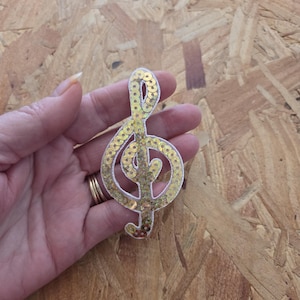 May include: A treble clef patch covered in shimmering gold sequins. The musical symbol is outlined with a white border, creating a distinct shape. The sequins are arranged to follow the curves of the clef, reflecting light across the surface.
