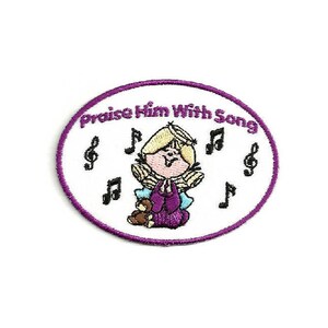 May include: Purple embroidered patch with a white background. The patch features a cartoon angel with blonde hair singing with a teddy bear. The text "Praise Him With Song" is written around the angel.