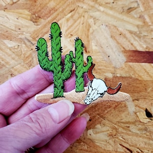 May include: Embroidered patch featuring a green cactus and a white bull skull with brown horns. The patch is on a tan background, with a desert theme. The cactus has black details.
