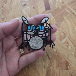 May include: An embroidered patch featuring a drum kit with a large silver bass drum, light pink tom-toms, and bright blue upper drums. The design is outlined in thick black thread and includes silver cymbals and black stands, creating a detailed musical instrument motif.