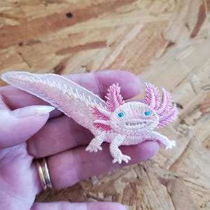May include: Embroidered axolotl patch in shades of pink and white, featuring blue eyes and detailed stitching. The patch depicts the aquatic salamander with its feathery gills and a smiling expression. Perfect for adding to clothing or accessories.