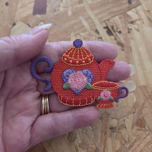 May include: An embroidered patch featuring a red teapot and a matching teacup. The teapot has a purple handle, a purple heart-shaped design with a pink flower center, and a purple lid knob. The teacup is red with a pink flower and purple handle.