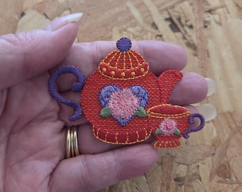 May include: An embroidered patch featuring a red teapot and a matching teacup. The teapot has a purple handle, a purple heart-shaped design with a pink flower center, and a purple lid knob. The teacup is red with a pink flower and purple handle.