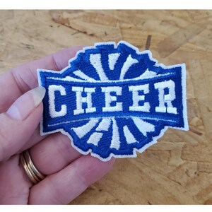 May include: Blue and white embroidered patch with the word "CHEER" in white lettering. The patch has a white and blue sunburst design.