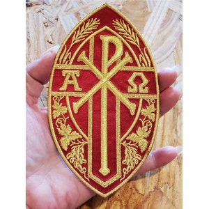 May include: A red oval patch with a gold embroidered Christian symbol, the Chi Rho, with the letters Alpha and Omega on either side. The symbol is surrounded by wheat and grapevine designs.