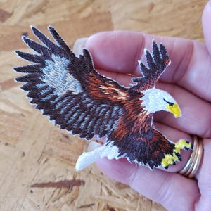 May include: Embroidered iron-on patch of a brown and white bald eagle with yellow talons and a yellow beak in flight.