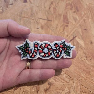 May include: Embroidered patch with the word "JOY" in white letters, featuring red and white candy cane designs and green holly leaves. The patch is a festive decoration for the holiday season.