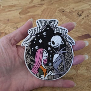 May include: Embroidered patch featuring Jack Skellington and Sally from The Nightmare Before Christmas. The patch is oval-shaped with a black background, snowflakes, and a white bow at the top. Sally has pink hair and an orange dress.