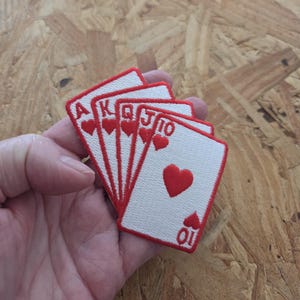 May include: A set of five embroidered patches shaped like playing cards, specifically the royal flush of hearts. Each white rectangular patch features a red border and red embroidery depicting the Ace, King, Queen, Jack, and Ten of hearts.